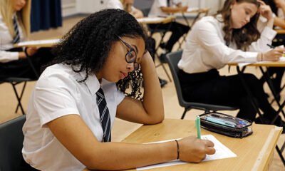 A female secondary school student sits an exam in a spacious school hall, the atmosphere calm and focused. She concentrates intently on her paper, pen in hand. The mid shot captures her quiet determination in the academic setting.