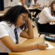 A female secondary school student sits an exam in a spacious school hall, the atmosphere calm and focused. She concentrates intently on her paper, pen in hand. The mid shot captures her quiet determination in the academic setting.